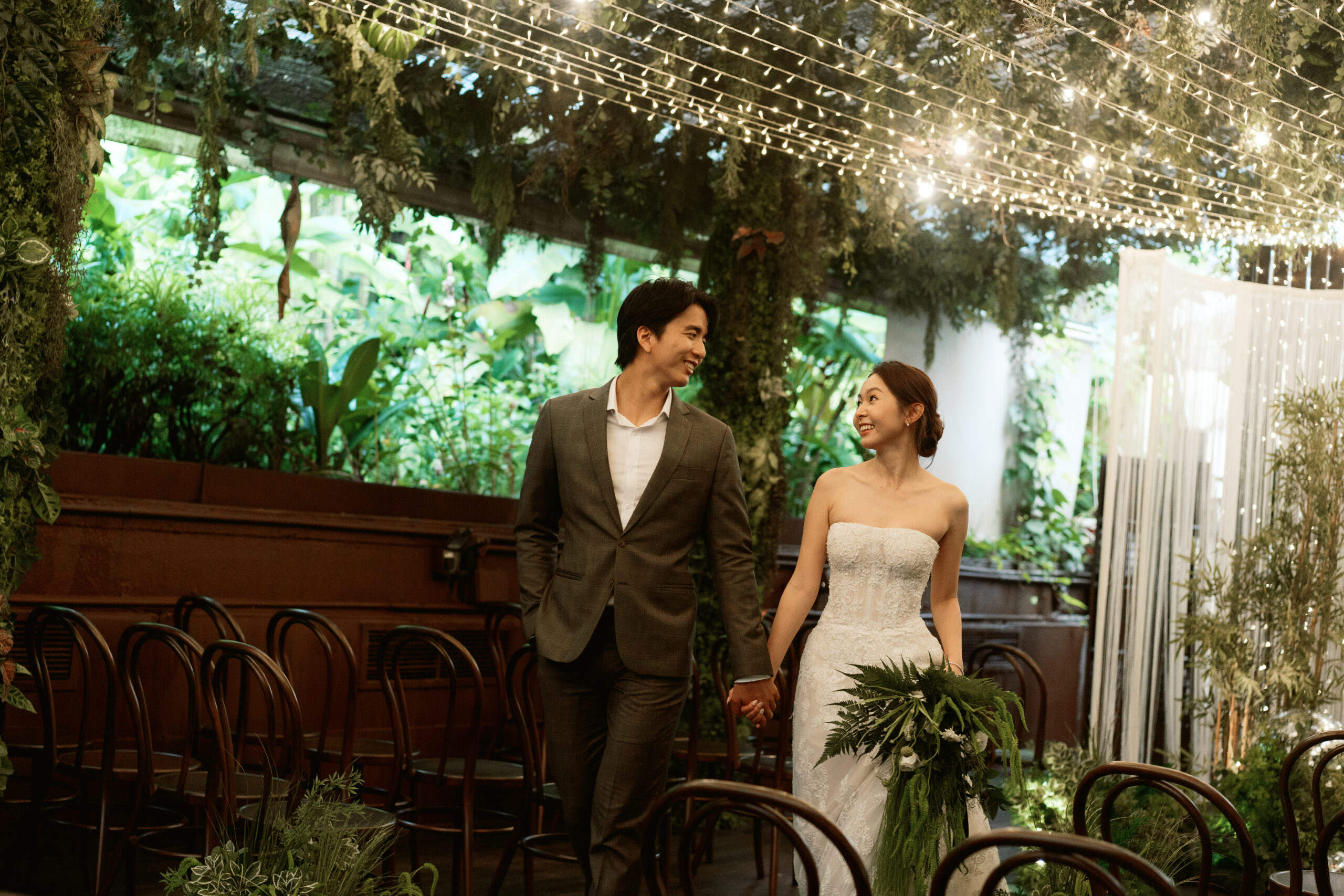 Couple walking down a glasshouse garden aisle under a canopy of fairy lights